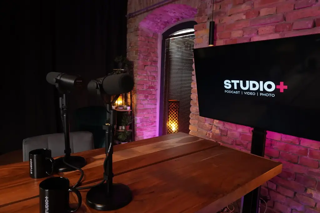 A STUDIO+ stúdió hosszú asztala mikrofonokkal, téglafallal és STUDIO+ feliratú képernyővel